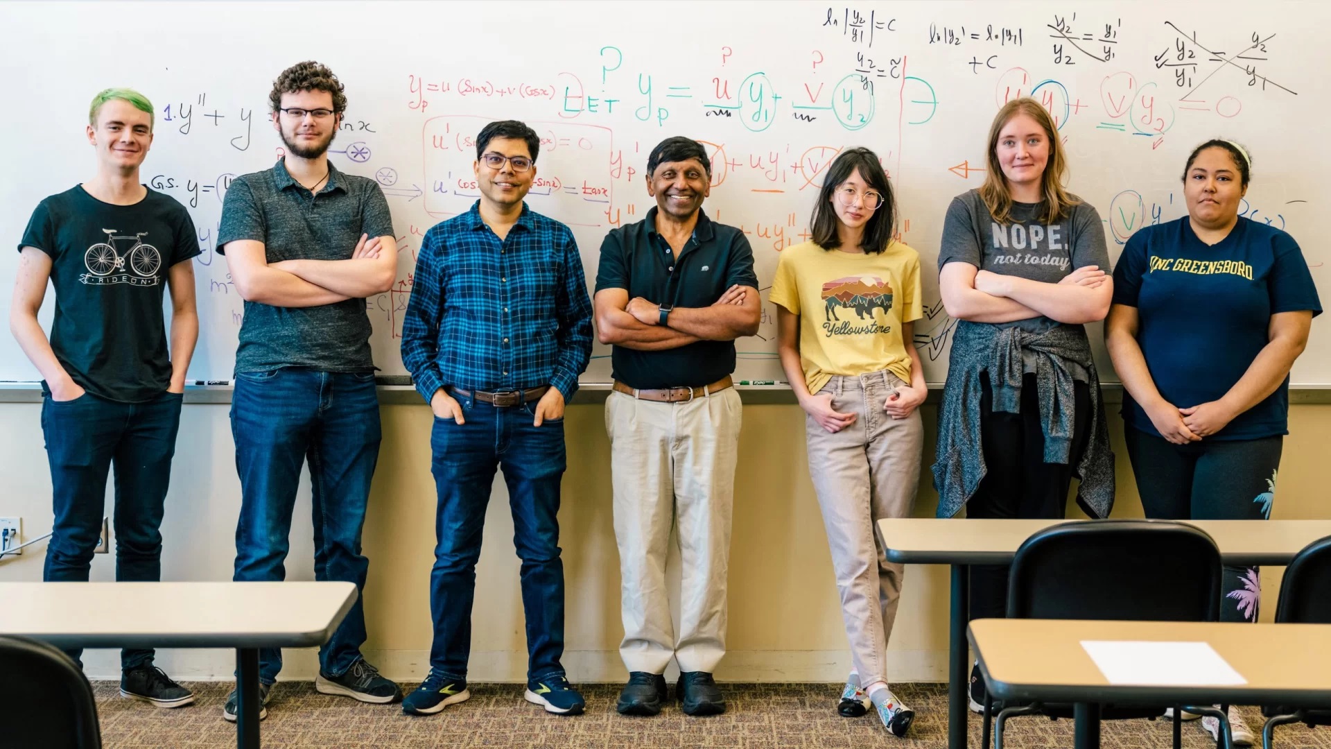Mathematics and Statistics professor Ratnasingham Shivaji poses in the middle of a group of students in front of a whiteboard with equations on it.